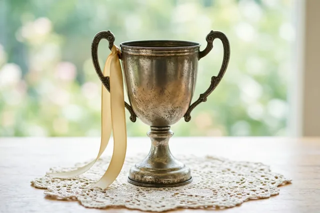 Een oude doffe metalen trofee met sierlijke handvatten en een crèmekleurig lint staat op een wit kanten kleedje op een houten tafel, met een wazig raam en groen op de achtergrond.