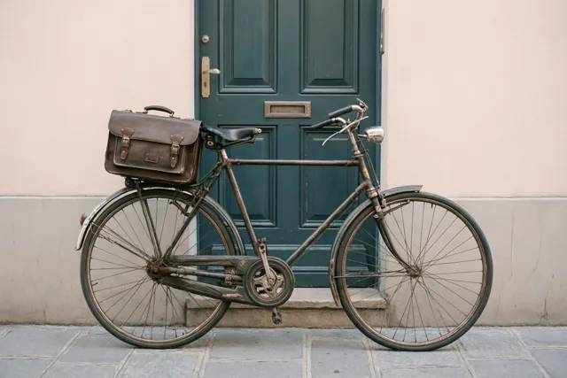 Een oude fiets met een bruine leren aktetas op de bagagedrager staat voor een donkergroene deur, tegen een lichtroze muur aan een grijze stenen stoep.