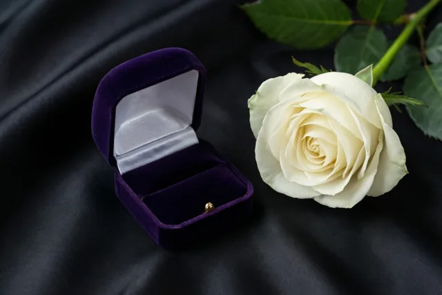 Een open paars fluwelen ringdoosje met een gouden ring staat naast één witte roos op zwarte zijde in zacht licht.