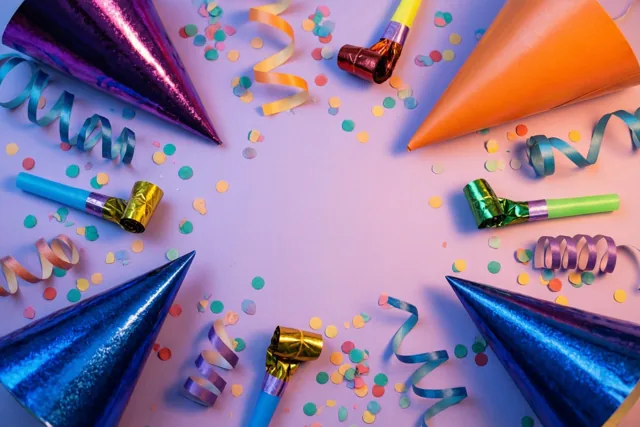 Van bovenaf zie je feesthoedjes, roltongen, confetti en gekrulde linten met matte en glanzende oppervlakken in een cirkel op een pastelpaarse achtergrond.