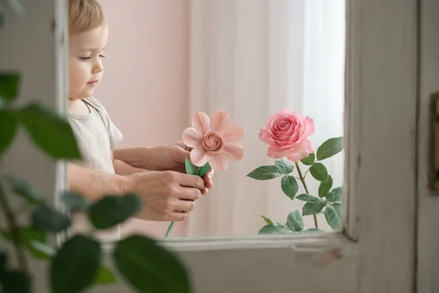Een jong kind houdt een zelfgemaakte papieren bloem vast die door volwassen handen wordt ondersteund, naast een echte roze roos op een vensterbank.
