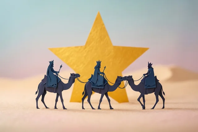 Drie uit papier geknipte silhouetten van koningen op kamelen trekken achter elkaar door een zanderige woestijn, met een grote gouden ster achter hen tegen een zacht verlopende lucht.