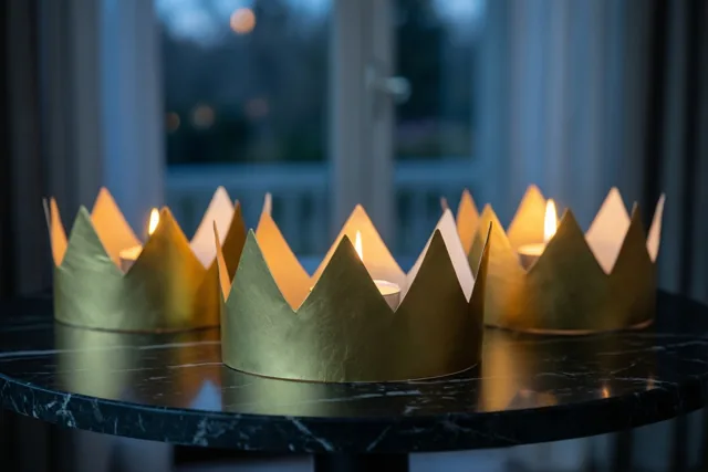 Drie gouden papieren kronen met elk een brandende kaars staan op een zwartmarmeren tafel, met een wazig raam op de achtergrond.