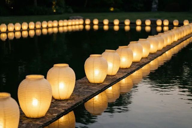 Rijen lichtgevende papieren lantaarns staan 's nachts langs een donkere vijver en weerspiegelen warm in het kalme water.