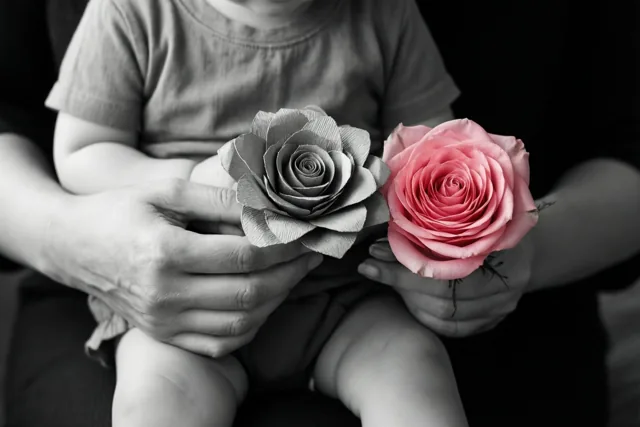 De handen van een moeder houden een door een kind gemaakte papieren roos naast een echte roze roos, de enige kleur in het verder grotendeels zwart-witte beeld.