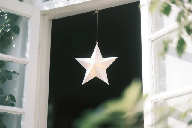 Een lichtgevende witte papieren ster hangt in een donker raam met een wit houten kader, met groene bladeren vooraan en opzij.