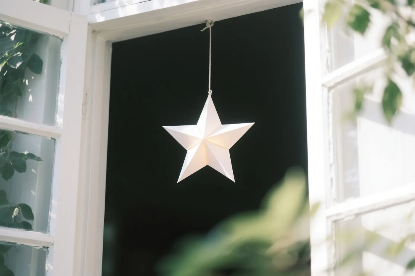 Een lichtgevende witte papieren ster hangt in een donker raam met een wit houten kader, met groene bladeren vooraan en opzij.