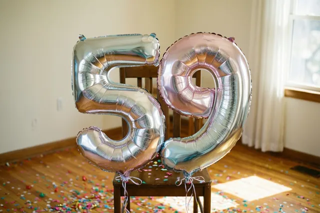 Twee glanzende folieballonnen vormen 59 in zilver en wisselende pasteltinten aan een houten stoel, met gekleurde confetti op de houten vloer.
