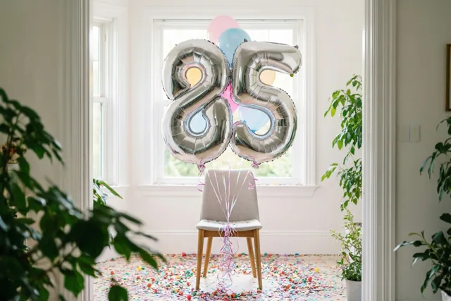 Zilveren en pastelkleurige cijferballonnen vormen 85 aan een stoel in een lichte kamer met witte muren, ramen, groene planten en confetti op de vloer.