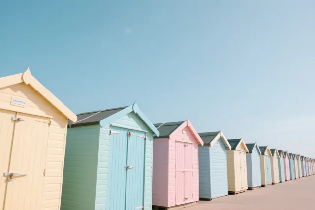 Een rij pastelkleurige strandhuisjes staat aan een zanderige kust onder een helderblauwe hemel.