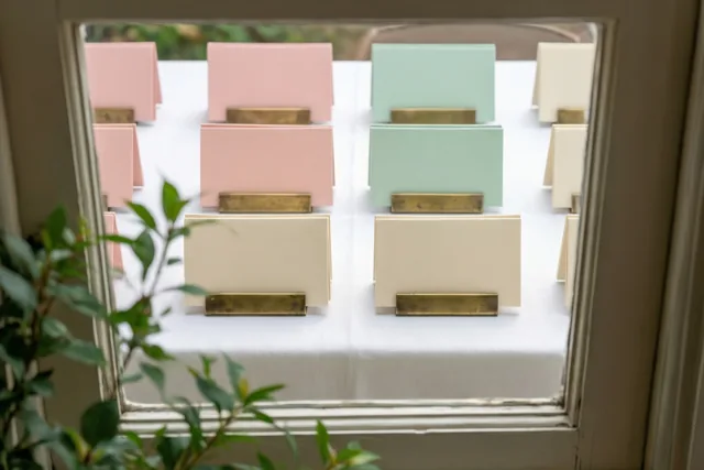 Tafelkaartjes in messing houders staan op een wit tafelkleed, gezien door een raamkader met groen op de voorgrond en zachte roze, mintgroene en crèmetinten.