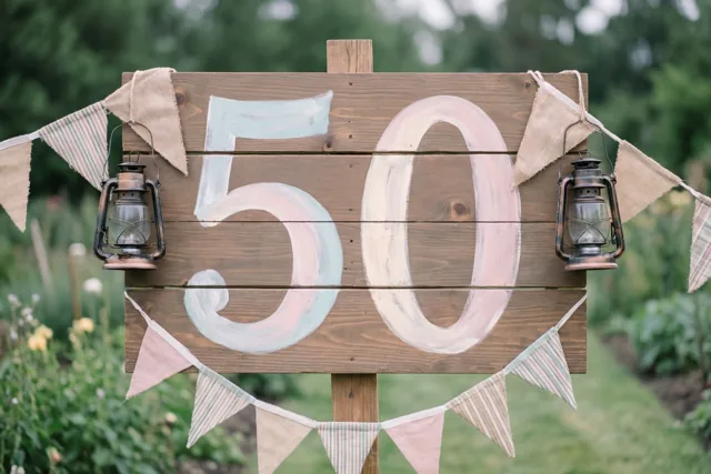 Een landelijk houten bord met 50 in pastelkleuren staat in een groene tuin met vlaggetjes en oude lantaarns.