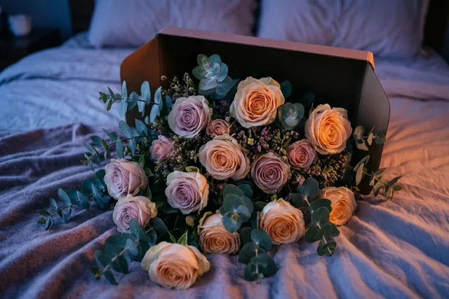 Een open bloemendoos ligt op een bed, met pastelkleurige rozen en eucalyptus die over een deken met structuur vallen.