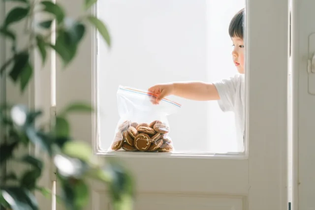 Een kinderhand reikt in een doorzichtige plastic zak pepernoten op een vensterbank, met groen op de voorgrond en een wit kozijn rond het raam.