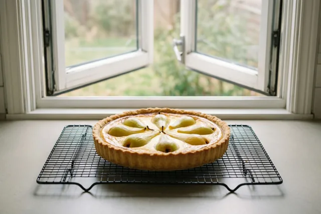 Een goudbruine perentaart met schijfjes peer in een cirkel staat op een zwart afkoelrooster op een witte vensterbank bij een open raam met groen tuinzicht.