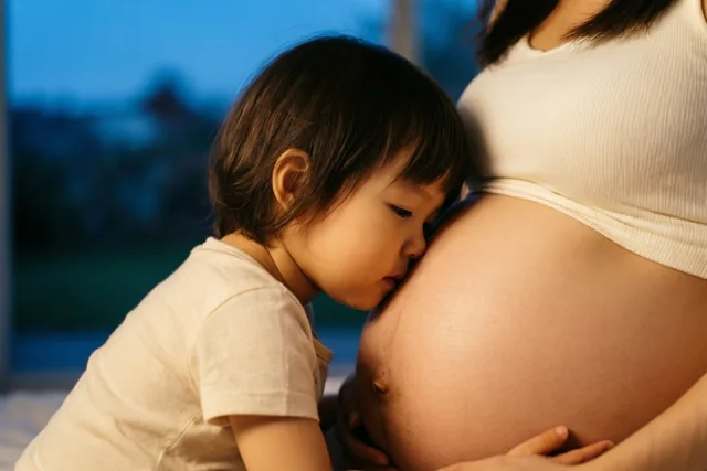 Van achter de peuter is te zien hoe ze haar wang tegen de ronde zwangere buik van haar mama legt, in warm licht tegen een blauwe achtergrond.