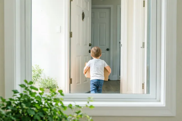 Een peuter in een blauwe jeans draagt een kleine kartonnen doos door een open voordeur, van achteren gezien vanuit een lichte kamer met witte muren, een wit raamkozijn en groene planten.