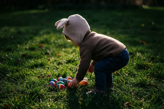 Een peuter met een muts met konijnenoren hurkt in het groene gras en reikt naar een beschilderd ei tussen andere eieren in warm zonlicht.