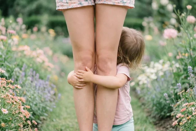 Een peuter met lichtbruin haar omhelst van achteren de benen van een vrouw in een tuin met kleurrijke bloemen en zacht daglicht.