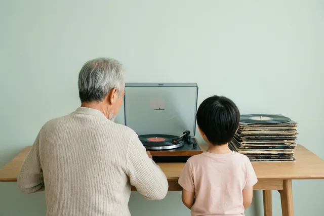 Een oudere man en een jong kind zitten met hun rug naar de kijker aan een houten tafel naar een platenspeler te luisteren, met platen ernaast en een lichtgroene muur erachter.
