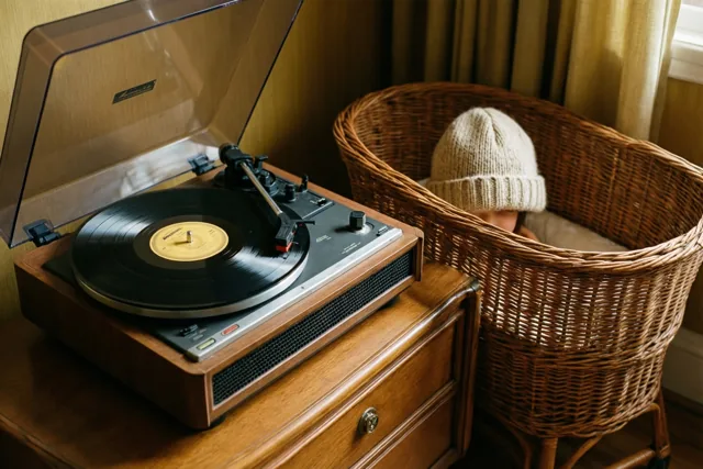 Een platenspeler in jaren-zestigstijl met een draaiende plaat staat op een houten ladekast naast een rieten wieg, in warm licht met beige en bruine tinten.