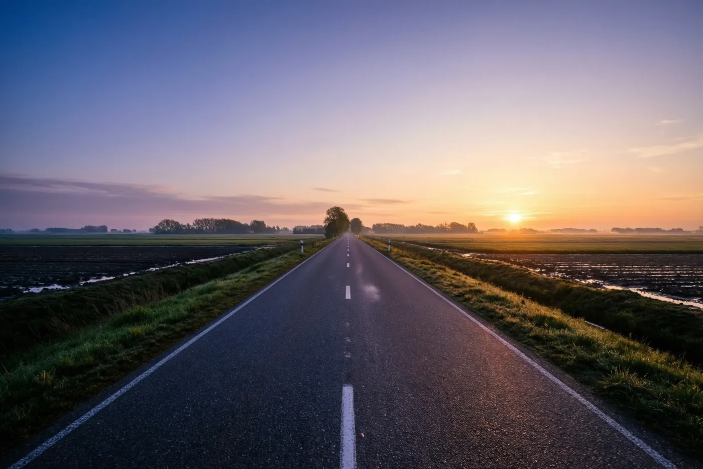 Een lange rechte asfaltweg loopt tussen grasbermen door vlakke polders met verre bomen, onder een hemel die bij zonsopgang van diepblauw naar oranje overgaat.