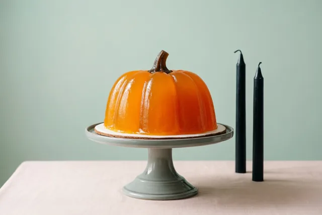 Een oranje geleipudding in pompoenvorm staat op een grijze taartstandaard op een lichtroze tafelkleed naast twee zwarte kaarsen tegen een lichtgroene muur.