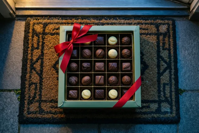 Een doos Belgische pralines met een rood lint staat op een deurmat met een patroon in warm licht.