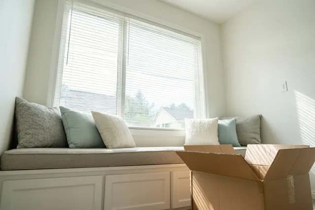 Een raambank met kussens staat in een lichte, pas ingerichte kamer met beige, witte en zacht blauwgrijze tinten.