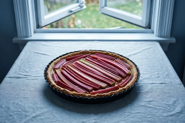Een rabarbertaart met rode stengels in een patroon staat op een blauw tafelkleed voor een open raam met natuurlijk licht.
