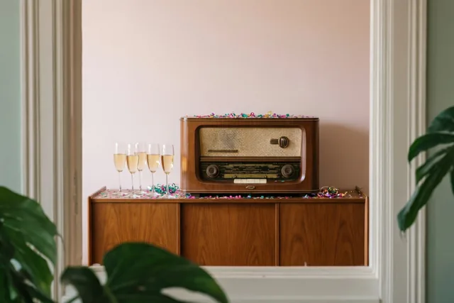 Door een deuropening is een houten radio op een dressoir te zien, met champagneglazen, confetti, monsterabladeren op de voorgrond en een zachtroze muur erachter.