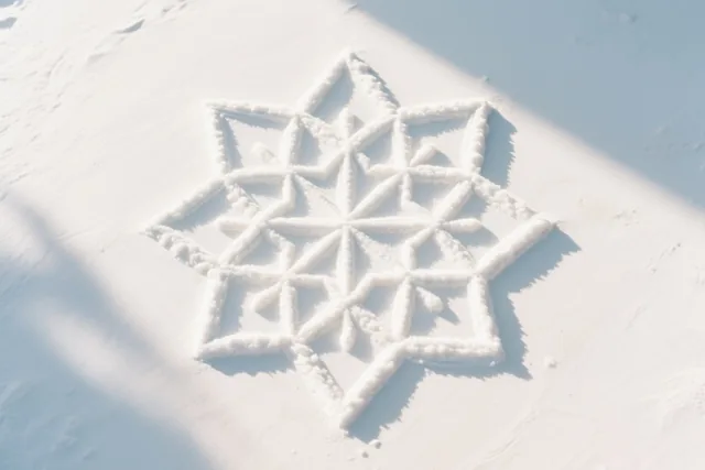 Van bovenaf is een geometrisch rangolipatroon in witte sneeuw te zien, dat op een sneeuwvlok lijkt en zachte schaduwen krijgt door het zonlicht.