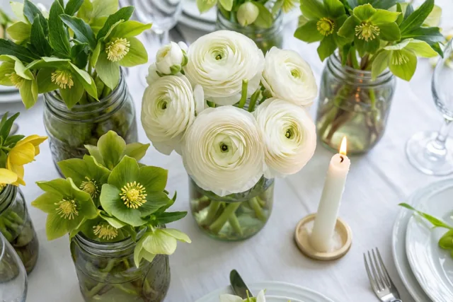 Witte ranonkels en groen nieskruid staan in glazen bokalen naast een brandende witte communiekaars op een tafel met een wit kleed, borden en bestek.