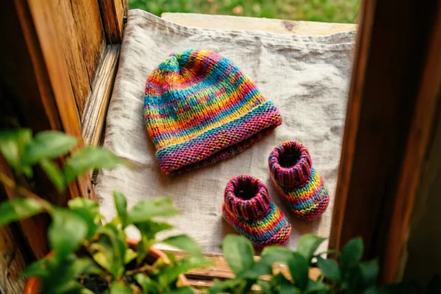 Een gebreid babymutsje en slofjes in regenboogkleuren liggen op beige linnen, van bovenaf gezien door een houten raamkozijn met houten panelen en groen gebladerte eromheen.