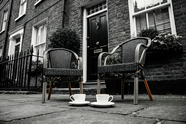 Twee rieten stoelen en twee witte koffiekoppen staan op het voetpad voor een bakstenen rijwoning in een overwegend zwart-witbeeld waarin de koppen en stoelpoten opvallen.