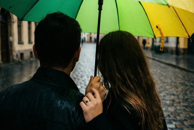 Een koppel staat met de rug naar de camera onder een groen-gele paraplu op een natte kasseistraat, waarbij de vrouw met een ring aan haar hand het handvat vasthoudt.
