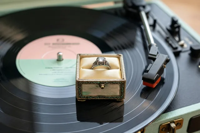Een klein vintage ringdoosje met een sierlijke ring staat op een draaiende vinylplaat op een retroplatenspeler, in gedempte pasteltinten.