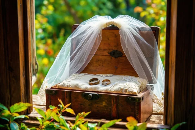 Op een vensterbank staat een oud houten doosje met twee versleten trouwringen op een kanten kussentje en een oude bruidssluier eroverheen, met groen op de achtergrond.