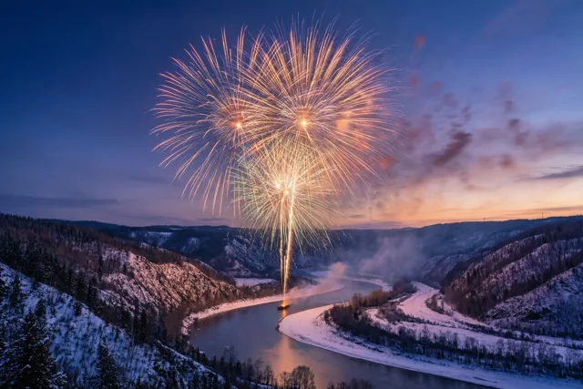 Oranje en geel vuurwerk barst los boven een rivier die door besneeuwde heuvels kronkelt onder een diepblauwe en paarse schemerhemel.