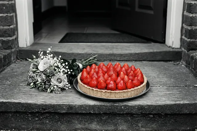 Een aardbeientaart en een klein boeket staan op een stenen drempel in een grotendeels zwart-witbeeld waarin de rode aardbeien opvallen.