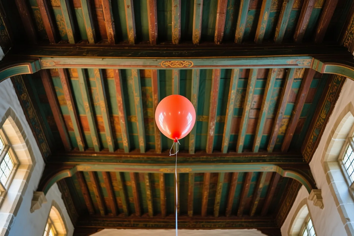 Een rode ballon zweeft onder een hoog houten plafond met groen en goud beschilderde balken in een oud Vlaams herenhuis met stenen muren en boogramen.