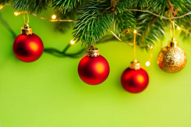 Rode en gouden kerstballen hangen aan een dennentak tegen een felgroene achtergrond, met warme lichtjes achter de ballen.