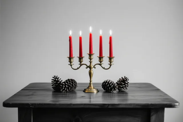 Een messing kandelaar met vijf brandende rode kaarsen staat op een donkere houten tafel met dennenappels, tegen een effen grijze achtergrond.