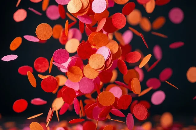 Papieren confetti in rode, oranje en roze tinten valt tegen een donkere achtergrond.