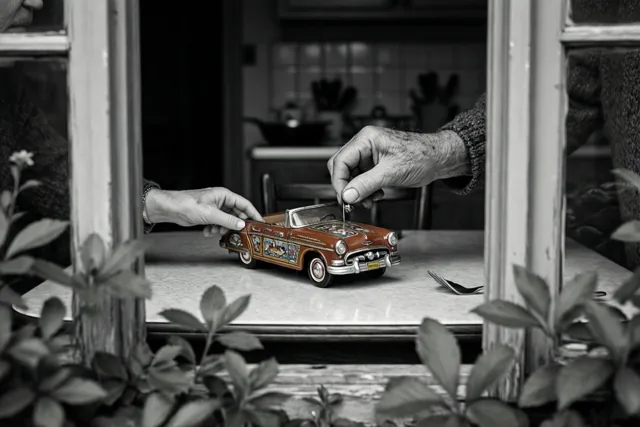Door een raamkozijn met groen op de voorgrond zijn de handen van een oudere persoon te zien die een blikken speelgoedauto op een keukentafel opwinden, in zwart-wit met alleen de carrosserie in rood.