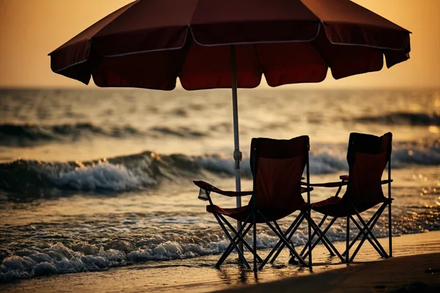 Een rode parasol en twee bijpassende klapstoelen staan aan de waterlijn van een zandstrand met zachte golven, in het warme goudkleurige licht van de zonsondergang.