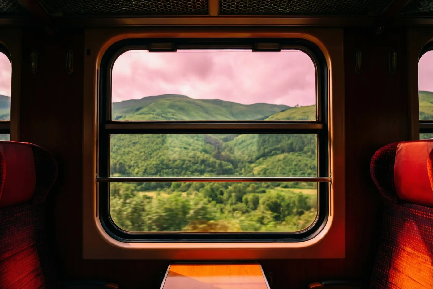 Vanuit een trein met rode stoelen met een patroon en houten afwerking zijn glooiende groene heuvels onder een roze hemel te zien.