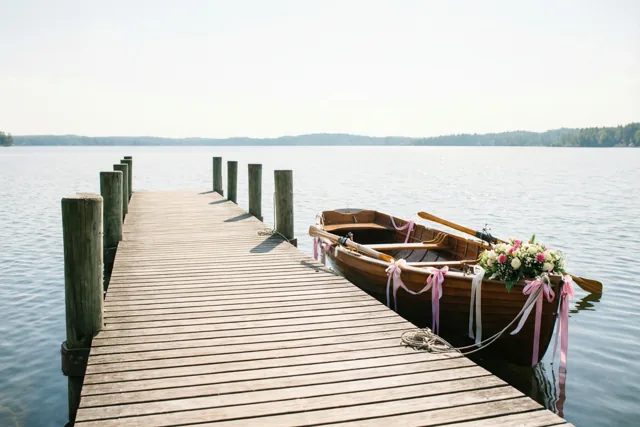 Een houten steiger loopt over rustig meerwater naar een vastgebonden roeiboot met roze en witte linten en een bloemstuk, met beboste oevers onder een heldere nevelige hemel.