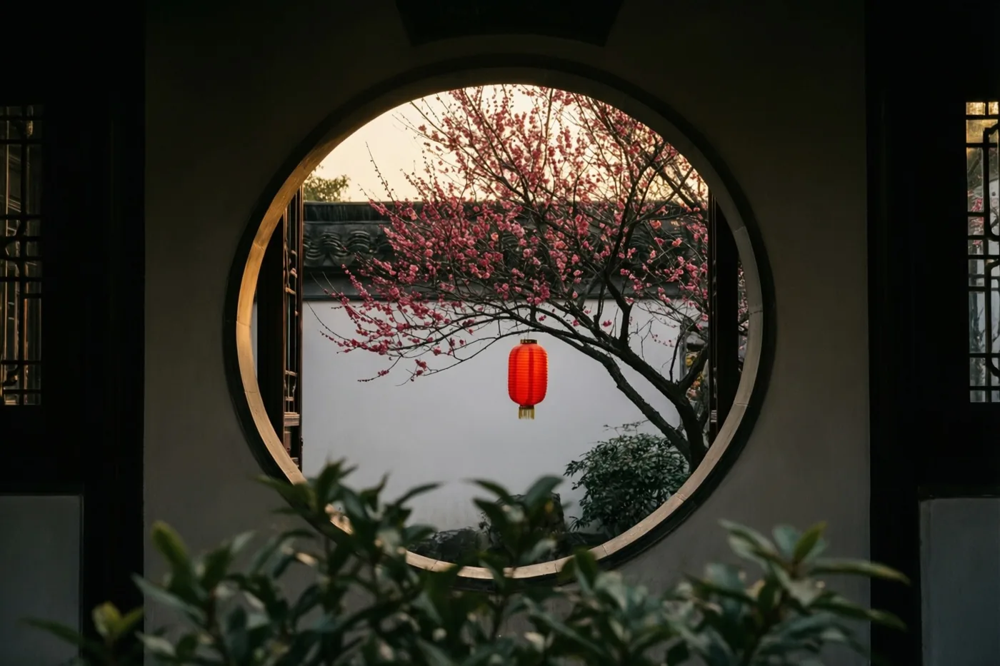 Een rond raam omlijst een tak roze pruimenbloesem en een hangende rode lantaarn voor een witte muur in de schemering, met groene bladeren op de voorgrond.