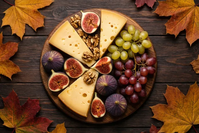 Een ronde houten kaasplank met plakjes kaas, hele en gehalveerde vijgen, groene en rode druiven en walnoten ligt op een donkere houten tafel tussen kleurrijke esdoornbladeren.
